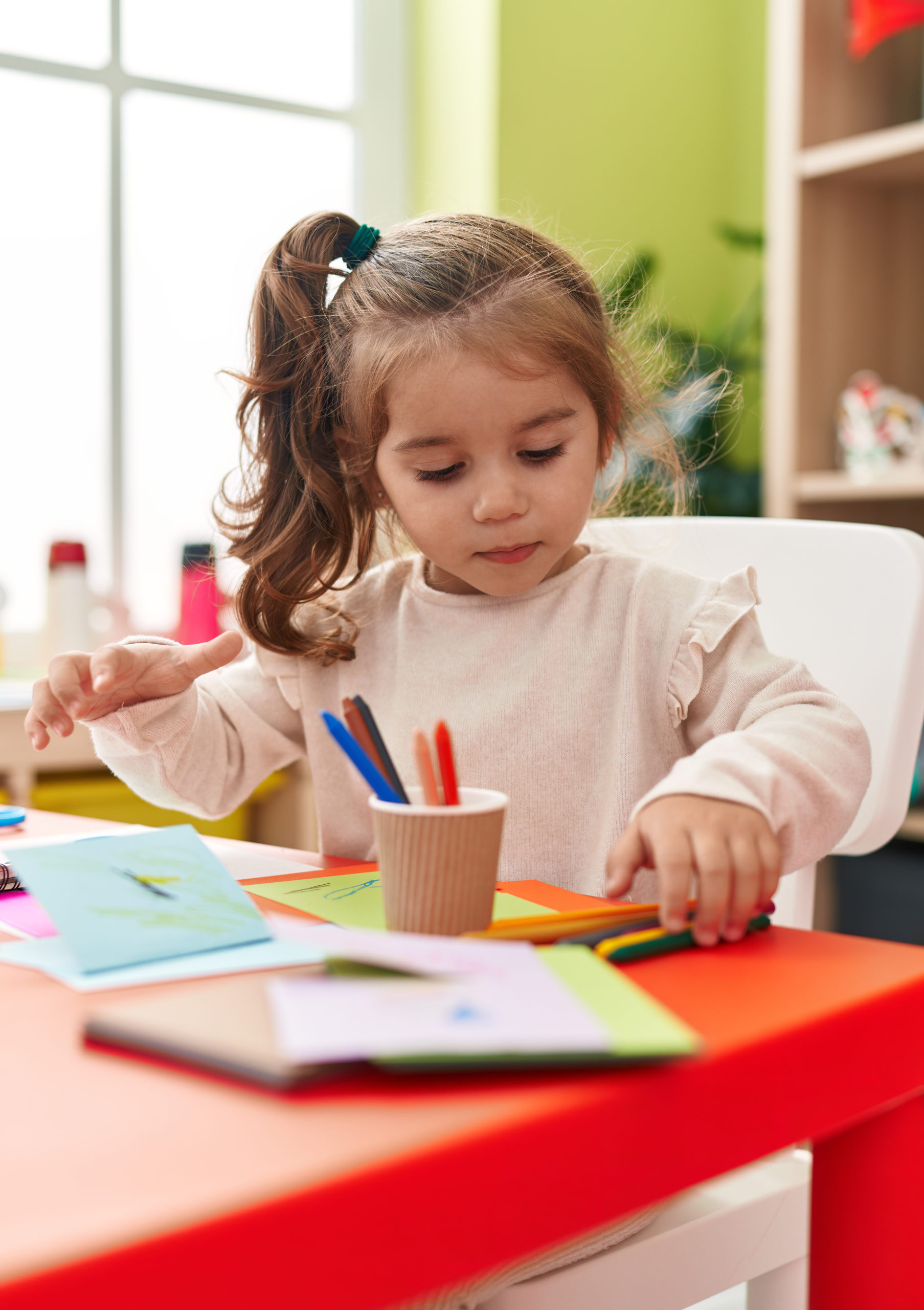 Adorable hispanic girl student sitting on table drawing on paper at kindergarten